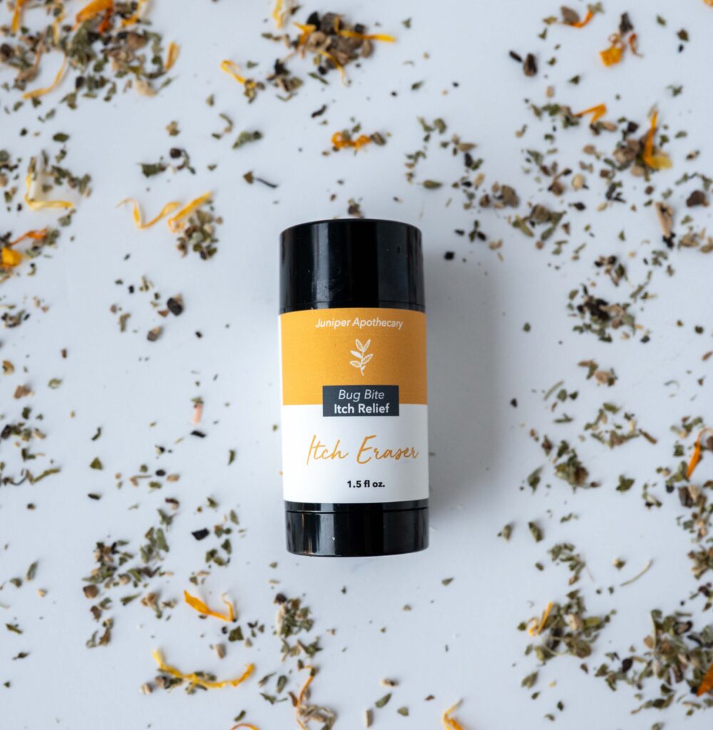 Juniper Apothecary Itch Eraser bug bite relief stick in a 1.5 ounce twist up tube, surrounded by scattered dried herbs including calendula petals, chickweed, and nettle, natural plant-based itch relief made with botanically infused olive oil