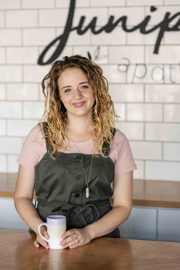 A smiling woman with curly hair stands behind a wooden counter, holding a coffee mug, against a backdrop of white tiles and a logo that reads 'Juniper'. She wears a green apron over a pink shirt.